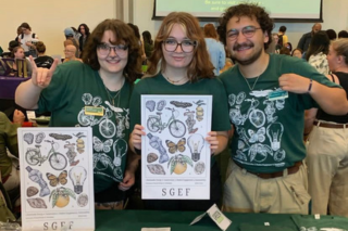 Three students wearing matching green tee shirts and holding up a poster with insects on it. 