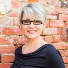 Woman wearing glasses and purple shirt smiling at camera with brick background