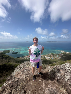 Student holding up double peace signs while on a high rock with blue water behind her. 