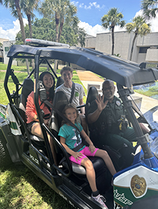 A group of four people, including three adults and one child, are seated in an off-road utility vehicle parked on grass. A uniformed officer, sitting in the front passenger seat, is smiling and waving. The scene is outdoors with palm trees and a building visible in the background under a bright blue sky.