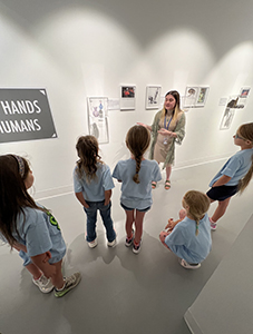 A group of young children wearing matching light blue shirts is gathered in an art gallery, attentively listening to a woman in a beige outfit who is leading the tour. The wall behind her displays various framed illustrations and a sign that partially reads, 'Hands Humans.' The space is well-lit with a clean, modern design.