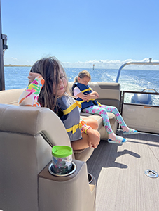 Two young children wearing life jackets are seated on a boat under a clear blue sky. The child in the foreground has long hair and colorful clothing, resting their arm on the back of the seat, while the child in the background is looking down, possibly at something in their hands. The boat is moving on calm water, with a distant shoreline visible in the background.